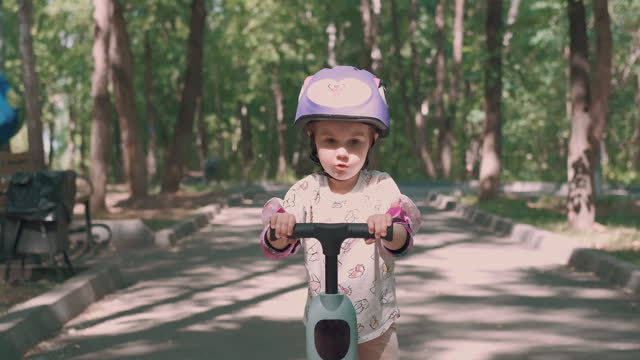 Mom teaches daughter to ride a scooter in the park in summer.