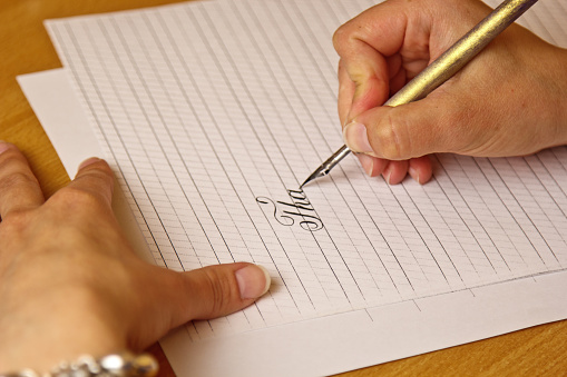 female hand writes with an ink pen on a white paper sheet with stripes. stationery on wooden desk close up top view. spelling lessons and caligraphy exercises.