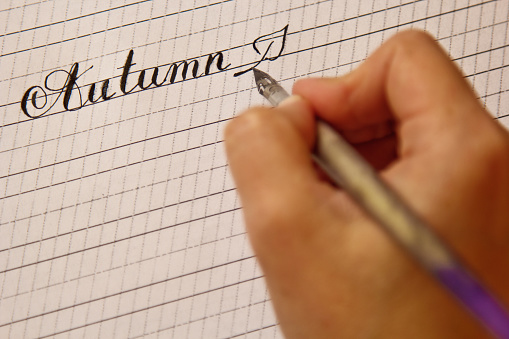 female hand writes with the inky pen the word autumn on a white paper sheet with stripes. stationery on desk close up top view. spelling lessons and caligraphy exercises. Template, layout, background.