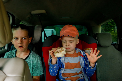 Two boys, brothers, a toddler and a schoolboy are sitting in a car. Children eat sweet donuts inside the car. Children in car seats. The car is parked. Snack on the trip.