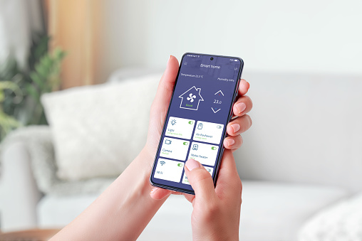 Woman's hand holding a mobile phone, showcasing a smart home app. Smart living concept. Decorated living room in background