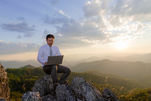 Man working outdoors with laptop sitting in mountains. Concept of remote work or freelancer lifestyle.Hiker tourist enjoying valley view sunset.