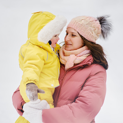 A mother woman holds a toddler baby in her arms, winter snow. Mom and son boy in warm clothes on the background of snowy nature. Kid aged one year eight months