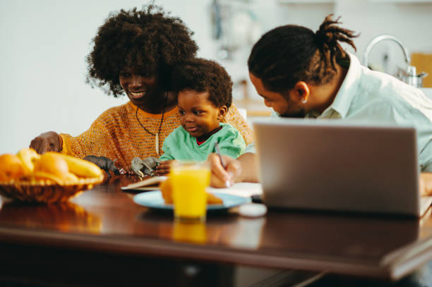 A happy multicultural family is sitting at home at the breakfast table and spending time together. An african american mother is holding her son in her lap and playing with him while her husband is sitting next to her and taking a break from remote work on a laptop. A happy family at home. morning time management for preschool for babies stock pictures, royalty-free photos & images