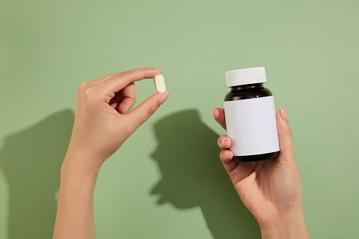 Two hand carrying a small white pill and a blank label medicine bottle. Pastel green background. Mockup of medicine brands
