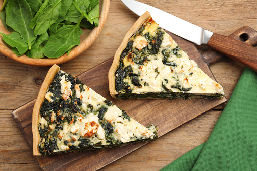 Pieces of delicious homemade quiche and fresh spinach leaves on wooden table, flat lay