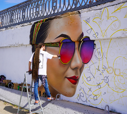 Young cheerful girl in a denim suit and a bright T-shirt is dancing with a sad face on the background of a wall with a yellow graffiti pattern