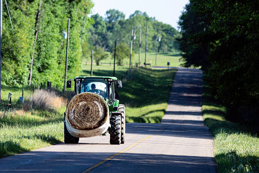Prattville, Alabama, USA:-April 18, 2023: Illustrative editorial image of a man hauling a round bale of hay from one field to another on a rural country road with a John Deere tractor.