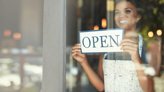 Small business, open sign and woman in cafe window for welcome, advertising and coffee shop promotion. Manager startup owner and excited and happy entrepreneur with opening poster for restaurant