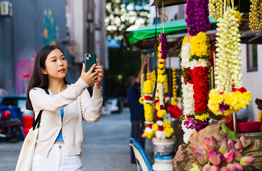 Asian woman taking photos using smart phone  hanged floral garlands in city.