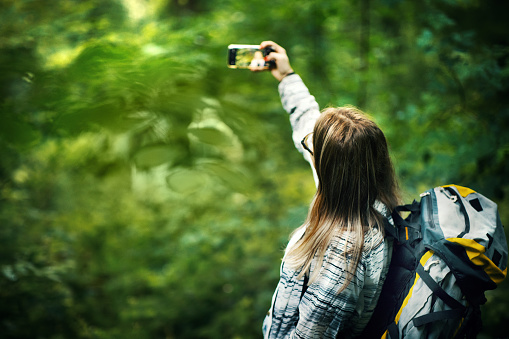 Closeup rear view of a mid 30's woman hiking through a forest and taking a selfie.