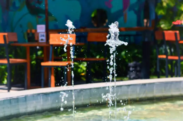 Close Up View Splashing Of Fountain In The Garden Pond With Table And Chairs Of The Garden Café