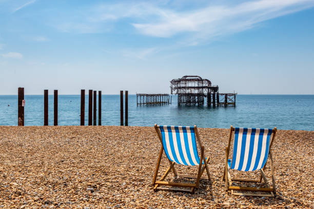 Two deck chairs on the pebble beach at Brighton on the Sussex coast, with the old West Pier behind A view of West Pier on Brighton Beach, with empty deck chairs in the foreground Hove stock pictures, royalty-free photos & images