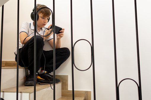 Boy with headphones playing games and surfing the internet on his smart phone sitting on the stairs at home.