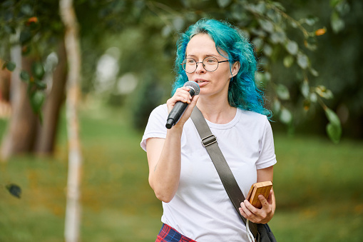 Young adult woman host of event in round glasses with turquoise dyed hair speaks into microphone at outdoor art exhibition, attractive female artist with bright appearance on green background