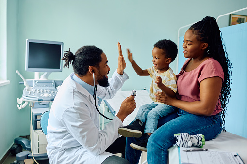 Happy African American doctor giving high five to a kid who is sitting on mother's lap during medical examination at pediatric clinic.