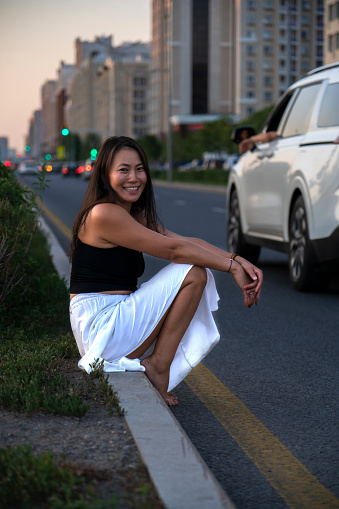 Asian girl with a smile on her face sits on the side of the road. Hitchhiking in summer.