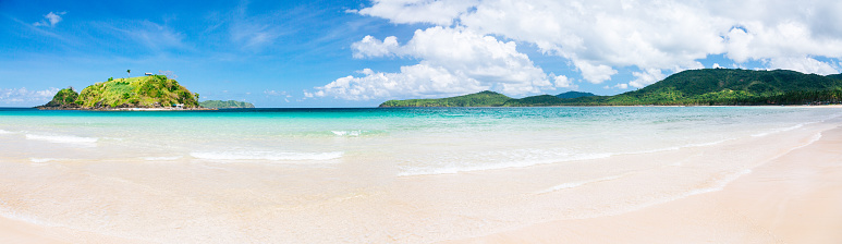 A panoramic view of Nacpan Beach, located on Palawan Island in the Philippines. The image captures a serene coastal landscape with a wide expanse of white sand stretching across the frame. Along the shoreline, there is a clear view of the calm and crystal-clear blue  sea meeting the sand. In the distance, a lush island can be seen on the horizon, covered with vibrant green vegetation. The sky above is blue, with a few scattered clouds adding a touch of texture. The overall scene exudes a sense of tranquility and natural beauty.