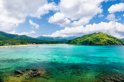 Aerial view of Palawan Island, focusing on a pristine blue lagoon. The lagoon's transparent waters offer a glimpse of visible coral reefs below. In the distance, a peninsula-hill covered with tropical plants stands prominently against the backdrop. The undulating terrain encompasses rolling hills and mountains, contributing to the overall landscape. Above, a clear blue sky with scattered clouds completes the scene. This unadorned photograph provides an unfiltered representation of the natural elements present in Palawan Island, allowing viewers to appreciate its serene beauty and unique topography.