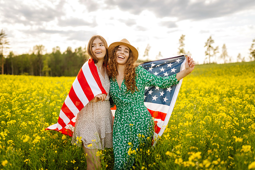 Two Young woman with american flag on blooming meadow. 4th of July. Independence Day. Patriotic holiday. USA flag fluttering in the wind.