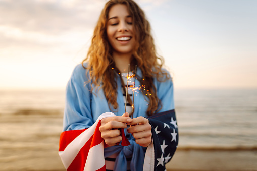 Young woman with American flag and sparklers on the beach. Patriotic holiday. USA celebrate 4th of July. Independence Day concept