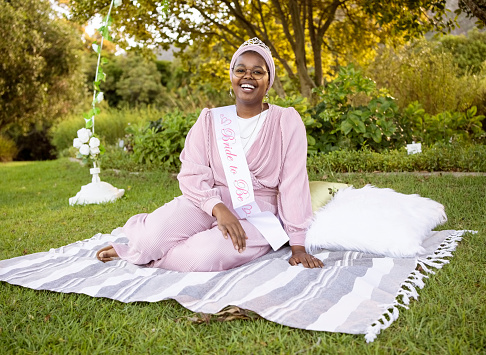 Smiling african woman wearing 'Bride to be' sash sitting on blanket at park for her bridal shower