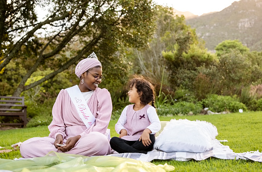 Smiling african woman wearing 'Bride to be' sash sitting with small girl on blanket at park for her bridal shower