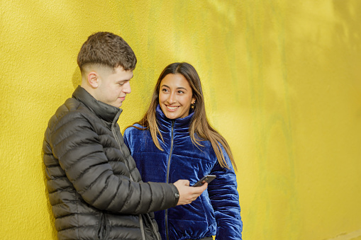 Couple of boy and girl leaning on a yellow wall watching the mobile phone with copy space.