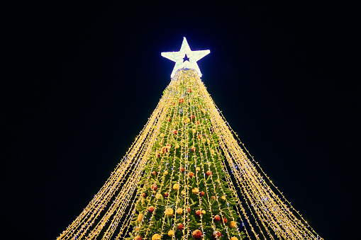 Christmas tree with yellow garlands, decorative bulbs and big white star topper at night blue sky background, outdoor holiday atmosphere. Festive Christmas tree with luminous yellow light garlands