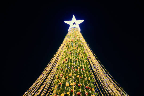 christmas tree with yellow garlands, decorative bulbs and big white star topper at night blue sky - celebration imagens e fotografias de stock