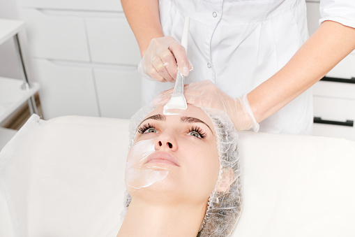 Beautician applies vitamins cream mask on woman face for rehydrate face skin, anti aging cosmetic procedure in beauty spa salon. Cosmetologist in gloves holds cosmetic brush for applying mask