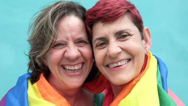 Happy Senior gay lesbian couple having fun wearing Lgbtq rainbow flag at pride parade - Family and love concept