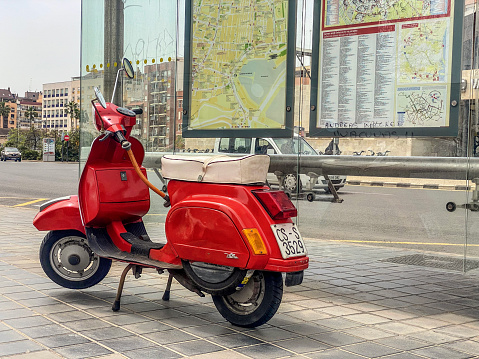Valencia - Spain - April 30, 2022: Side view of red motor scooter parked next to bus stop in the city of Valencia, Spain. This is a very used alternative to cars to move quickly and efficiently all around the city