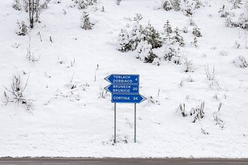 View of a roadsign by the road with directions to Cortina d'Ampezzo, Brunico and Dobbiaco, in a snowy winter day; Dolomites, Italy