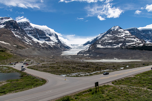 High angle view of the Icefields Parkway with in the background the Columbia Icefield in Jasper National Park, AB, Canada against a white clouded blue sky