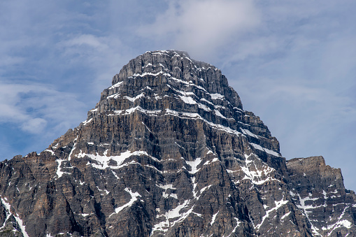 Close up of the partly snow covered top of the White Pyramid mountain in the Canadian Rockies of Banff National Park against a white clouded blue sky