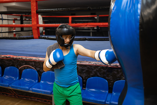 Young Chinese man going for boxing training. In this photo he is practicing punches on punching bag next to the boxing ring.