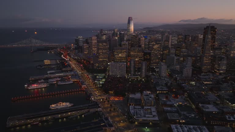 Fly above metropolis at dusk. Aerial footage of coastal road and piers along bank. Group of downtown skyscrapers with lighted windows.