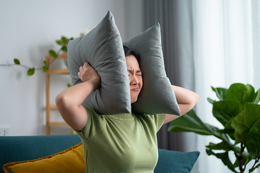 Asian woman feel boring have a problems with noise covering ears with pillows sitting on sofa in living room at home.