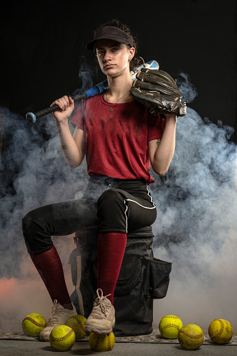 Athletic fast pitch softball player in red and black uniform sits on coaches bucket with foggy background.