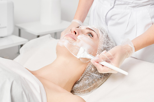 Beautician applies vitamins cream mask on woman face for rehydrate face skin, anti aging cosmetic procedure in beauty spa salon. Cosmetologist in gloves holds cosmetic brush for applying mask