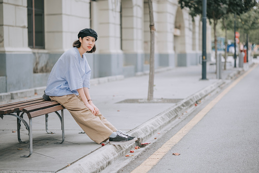 Asian Chinese young woman sitting on bench side walk waiting looking away