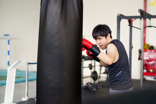 Active Asian sportsman practice an indoor boxing at the boxing gym, sportive man practice boxing alone with a sandbag alone. Fitness portrait of good looking Asian sportsman posing for photography then punching and kicking at boxing sandbag in the gym.