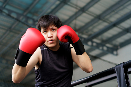 Active Asian sportsman practice an indoor boxing at the boxing gym, sportive man practice boxing alone with a sandbag alone. Fitness portrait of good looking Asian sportsman posing for photography then punching and kicking at boxing sandbag in the gym.