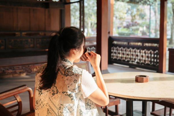 une femme asiatique boit du café et s’amuse lorsqu’elle est assise dans sa maison joglo (maison traditionnelle de java) - back against the wall photos et images de collection