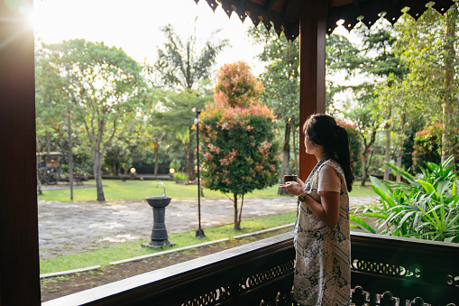asian woman drink coffee and enjoy when standing at her  joglo house (a type of traditional vernacular house of the Javanese people) (Javanese omah)