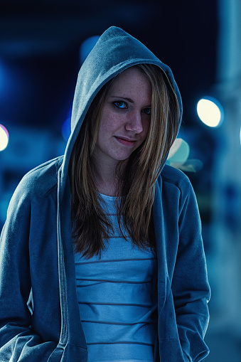 Portrait of a teenage girl wearing a gray hooded sweat shirt in a night time street scene, with a troubled expression on her face