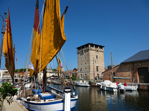 Cervia, Ravenna province, Italy June 17, 2023. Panoramic view of the canal and the St. Michael Tower circa 1700 once defended the city on the Adriatic Sea. The old seafaring boats with the original colored sails and coats of arms of fishing families.