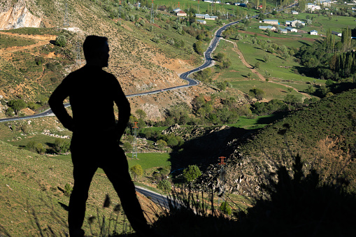 Silhouette view of man looking from the summit to the town surrounded by high mountains. The curved highway descending from the mountain to the valley is seen. In the spring season, the soil is green. Shot with a full-frame camera in daylight.