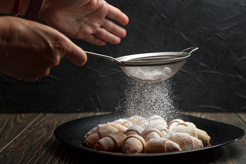 Womens hands sprinkle powdered sugar through a sieve on freshly baked rouge French croissants. Homemade baking concept